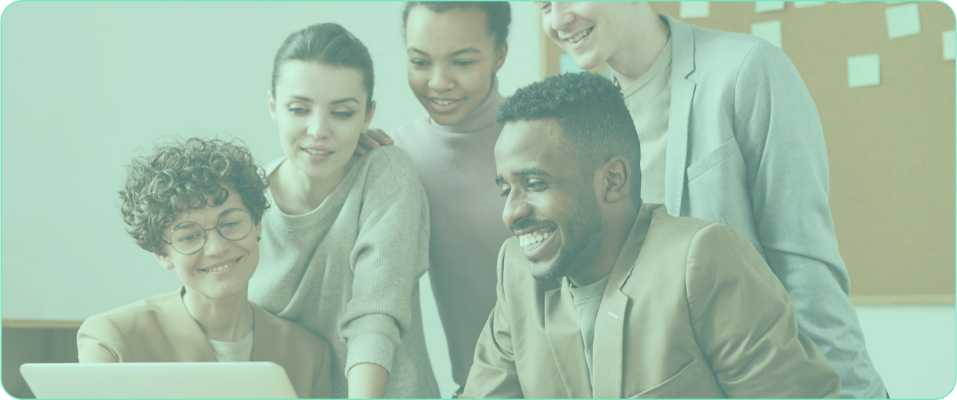 Group of diverse colleagues gathered around a laptop, smiling and collaborating, against an office background with a corkboard.